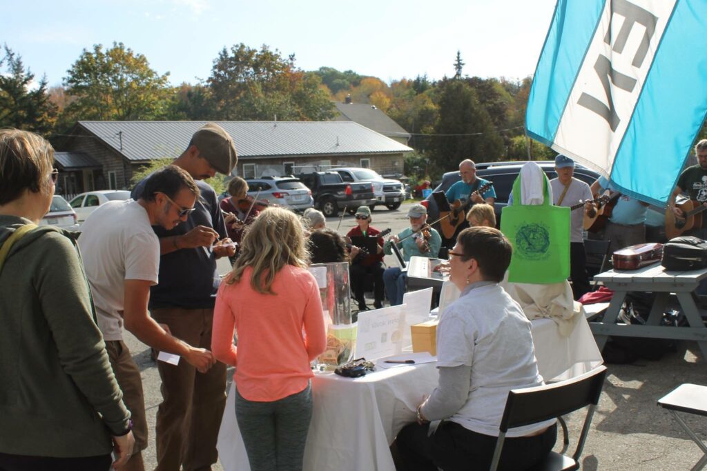 An image of people seated and standing around a table, gathered around outside on a nice sunny day. Musicians are playing instruments in the background, there is the suggestion of a flag that waves and reads OPEN.