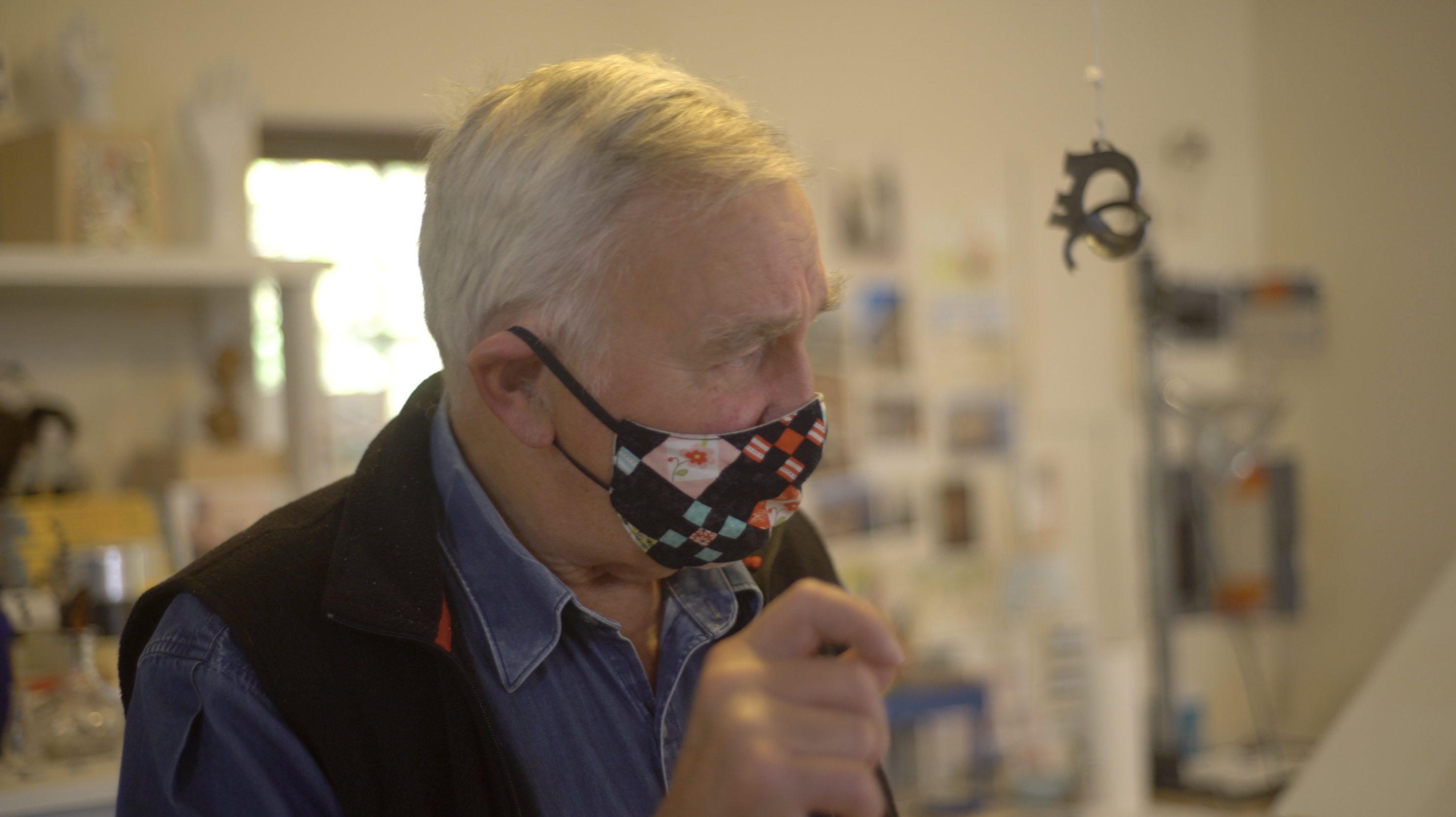 An image of a man wearing a cloth face mask and white hair, holding his right hand up as to suggest holding a writing or artistic implement.