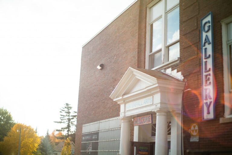 The Waterfall Arts building, a square brick building with Italianate white columns and portico, next to a sign that reads GALLERY oriented vertically and some sun flare throughout the image.
