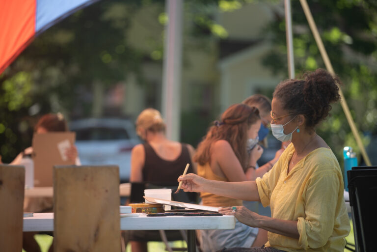 An image of a woman wearing a mask and a yellow shirt sitting at a table outdoors painting..